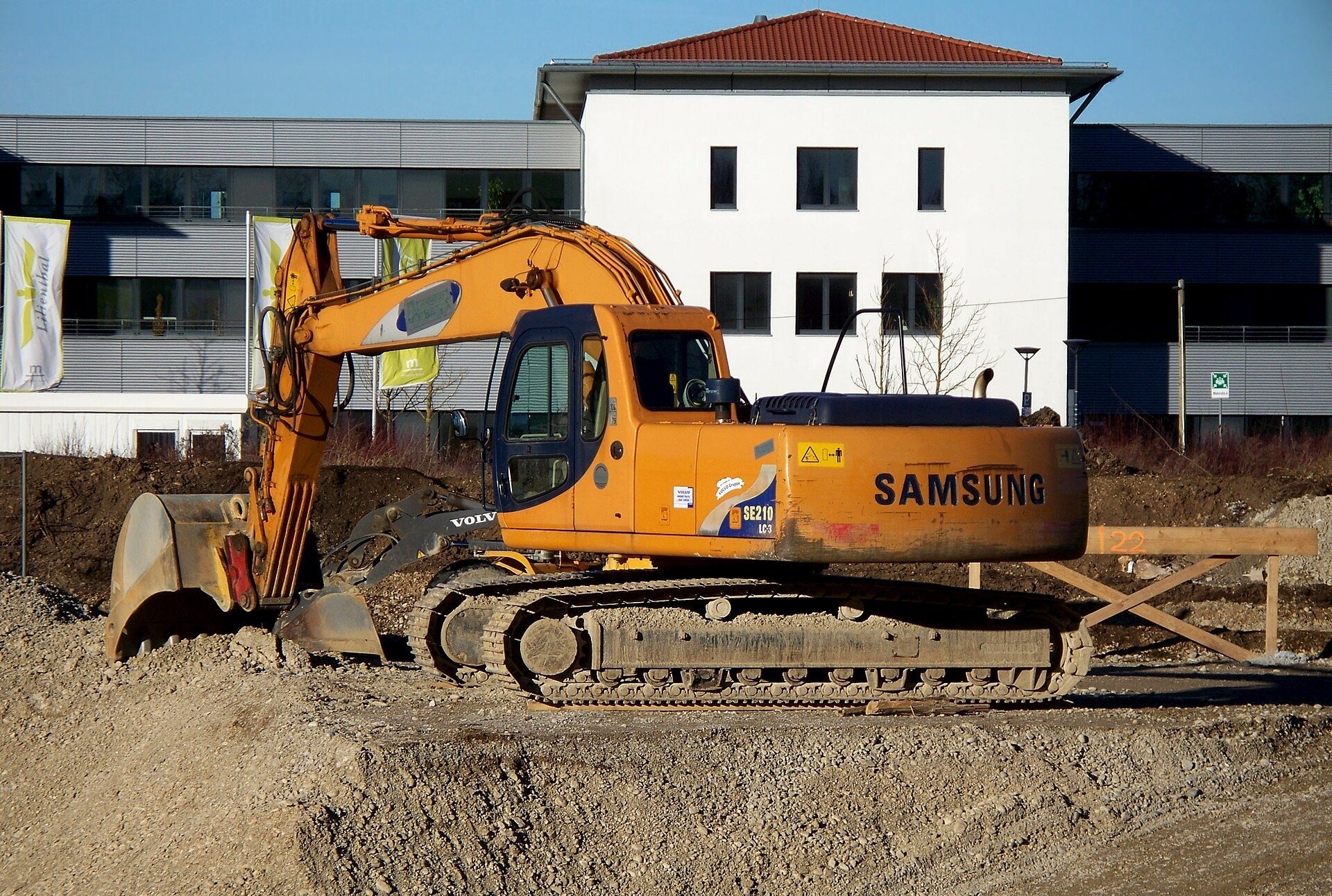 A Samsung excavator at a construction site, showing hydraulic cylinders on the boom and arm