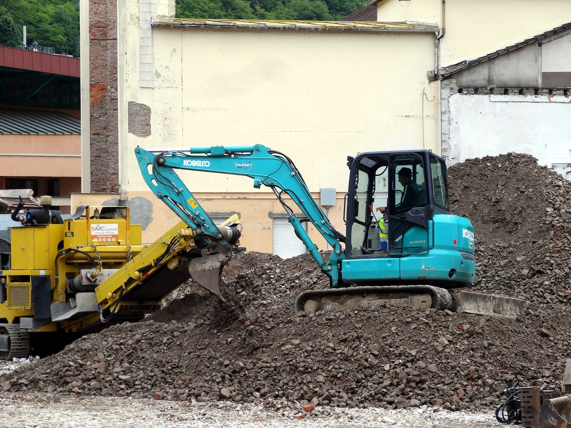 An operator visible inside the cab of a Kobelco compact excavator while digging