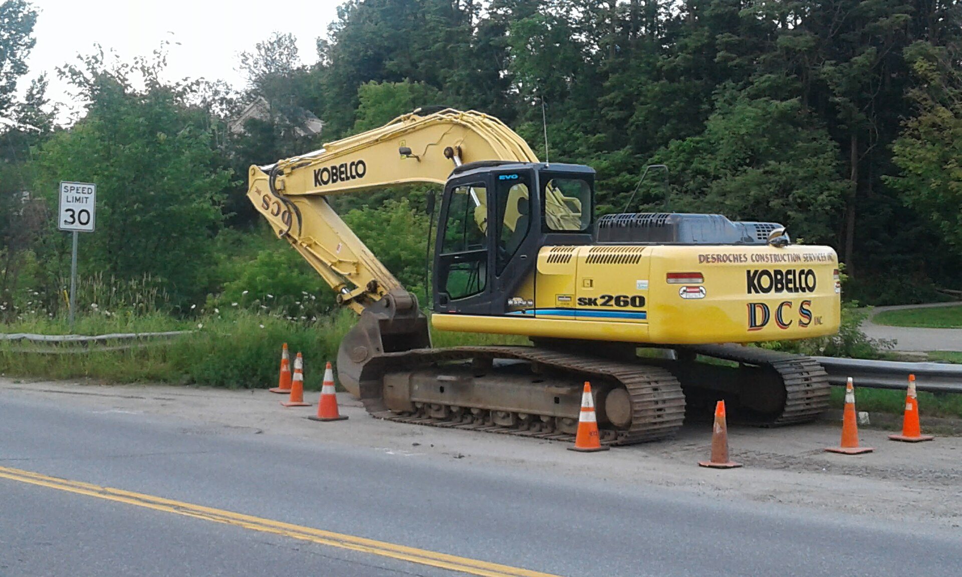A large blue Kobelco excavator parked at a job site, showing its long boom, arm, and bucket