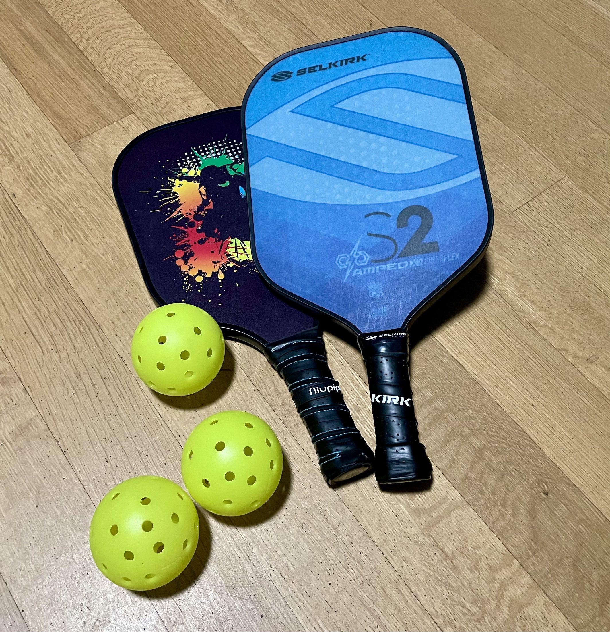 Three colorful pickleball balls and two paddles resting on a wooden surface, showing the perforated ball design